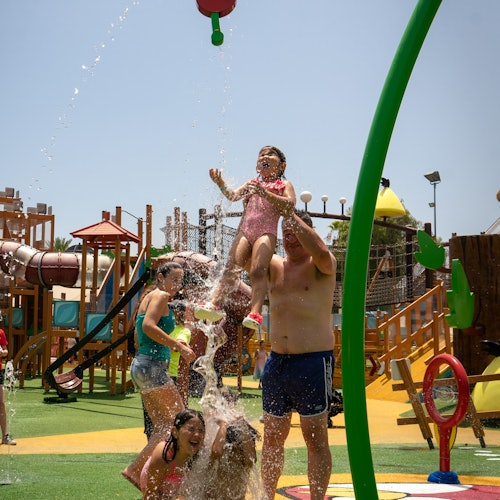 在一个充满活力的游乐场中，儿童和成年人在喷水下嬉戏，游乐场配备了攀爬设施和滑梯。