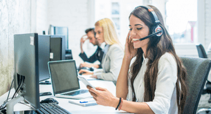 A woman at a row of computers talks on a headset while holding a cell phone.