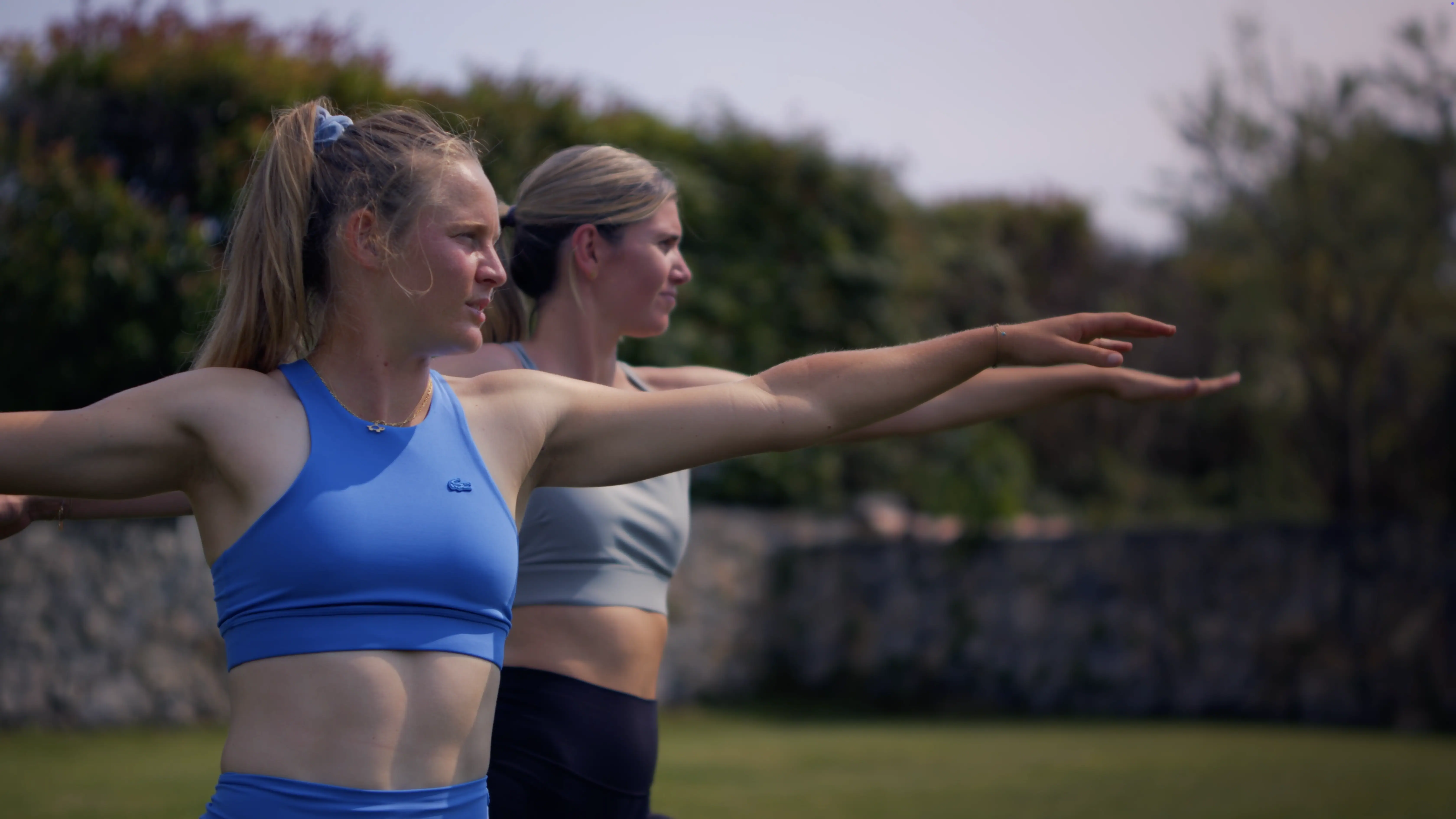 Two women practice yoga poses in a grassy outdoor setting, each extending their arms forward with focus and balance, surrounded by lush greenery and a stone wall in the background, highlighting fitness and outdoor exercise.