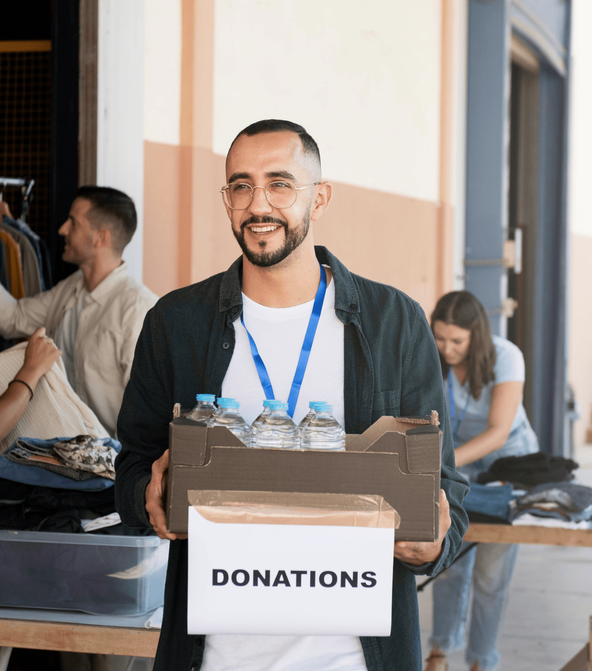 A smiling man holds a donations box at a busy collection event, with people and bags in the background.