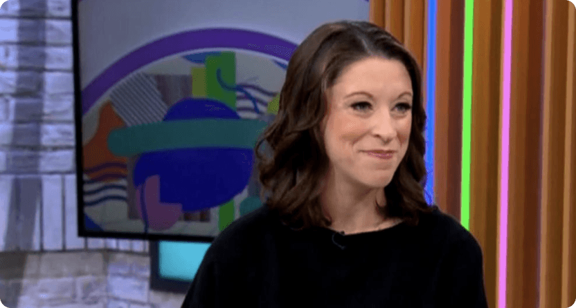 A woman with dark, curly hair wearing a black top, standing in front of a colorful, abstract background, with a warm, friendly expression on her face, suggesting a professional setting such as a television studio.