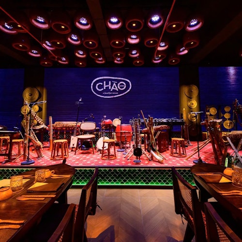Sebuah panggung dengan alat musik yang dikelilingi oleh meja, kursi, dan logo bertuliskan "CHÁO" pada latar belakang biru, di bawah pencahayaan dramatis.