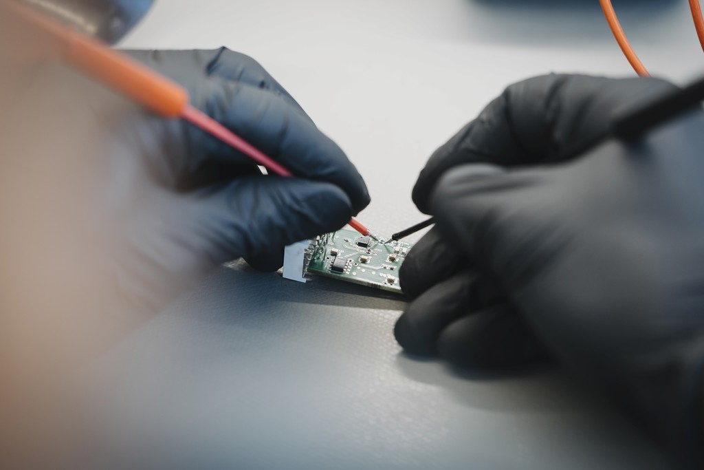 A close-up of two hands in black gloves performing delicate, precise work on a tiny circuit board with a fine tool.