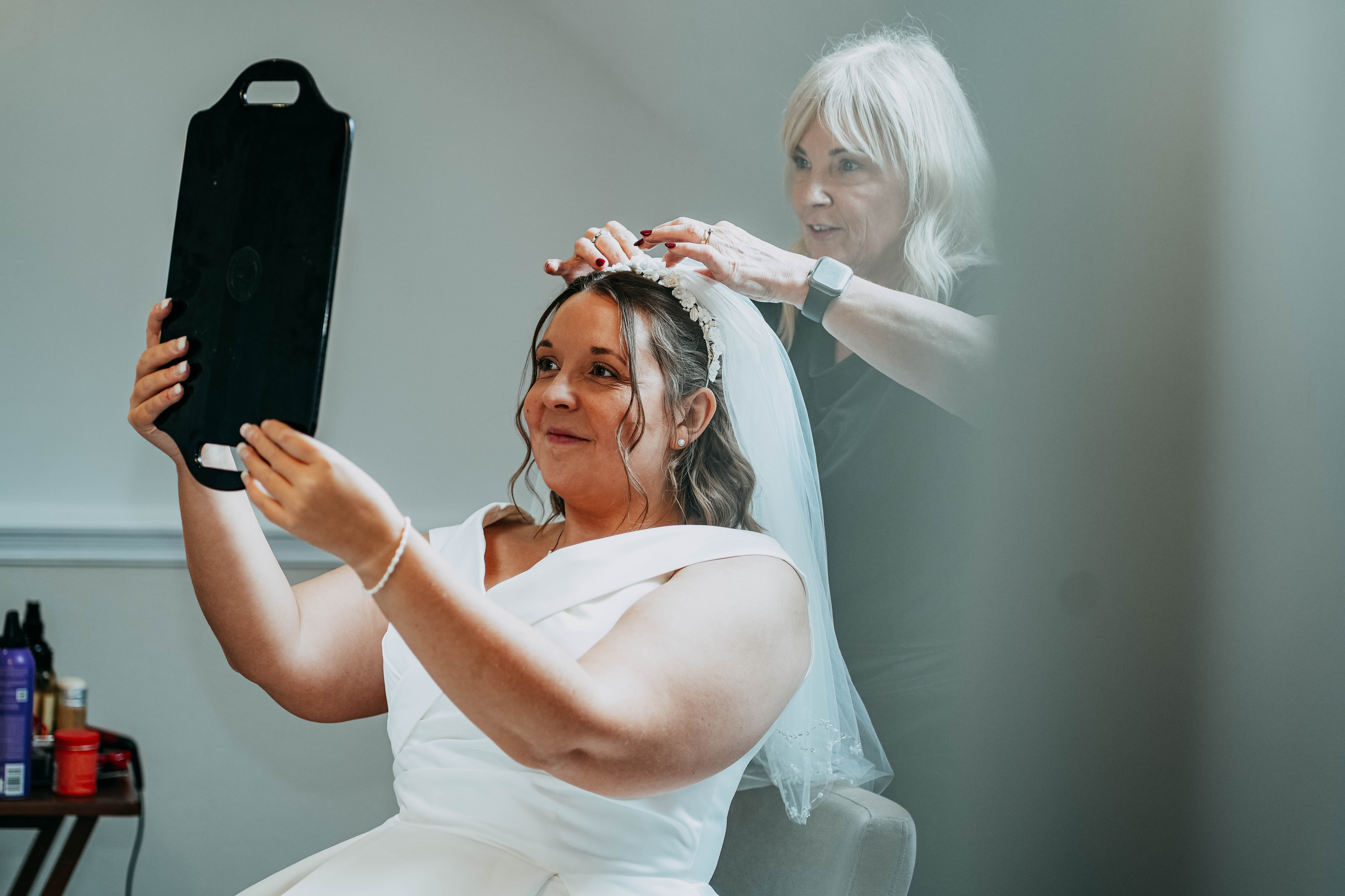 The bride in her white wedding gown smiles as she looks into a handheld mirror while another woman adjusts the tiara and veil on her head.