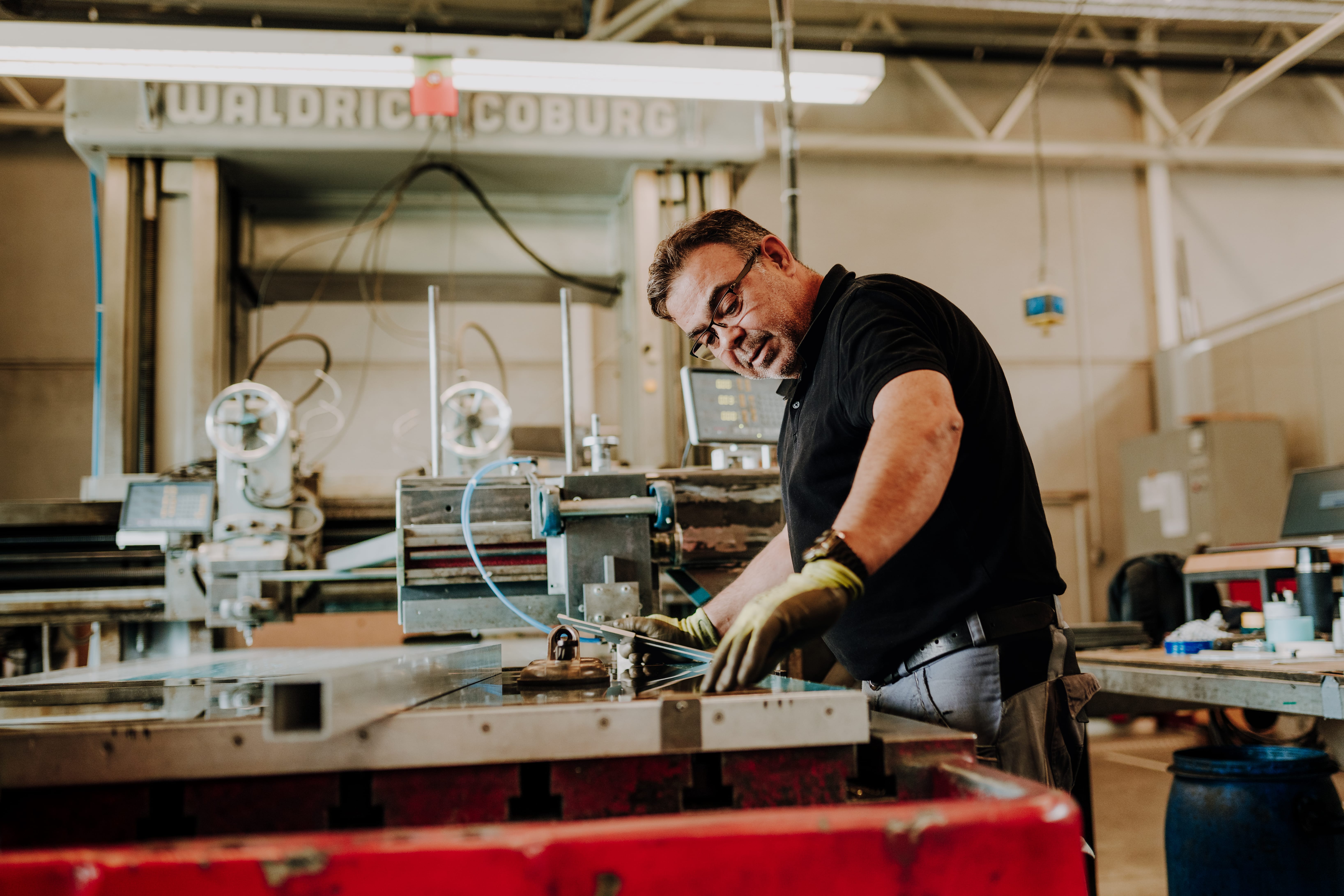 Man in glasses works on a machine in a workshop. "WALDRICH COBURG" machine is in the background.