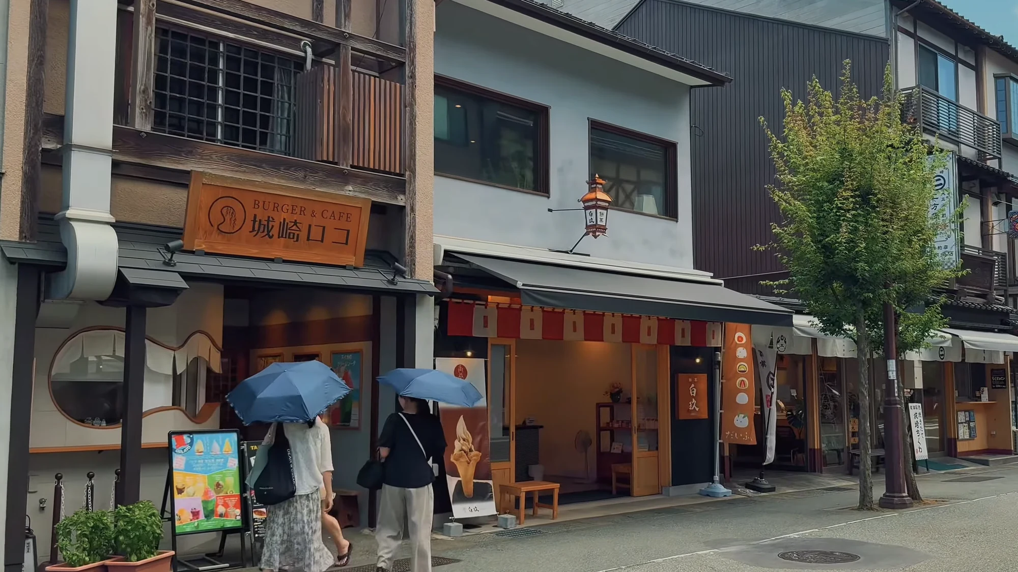 Two visitors sheltering under blue umbrellas outside a cafe on the Kinosaki main street.