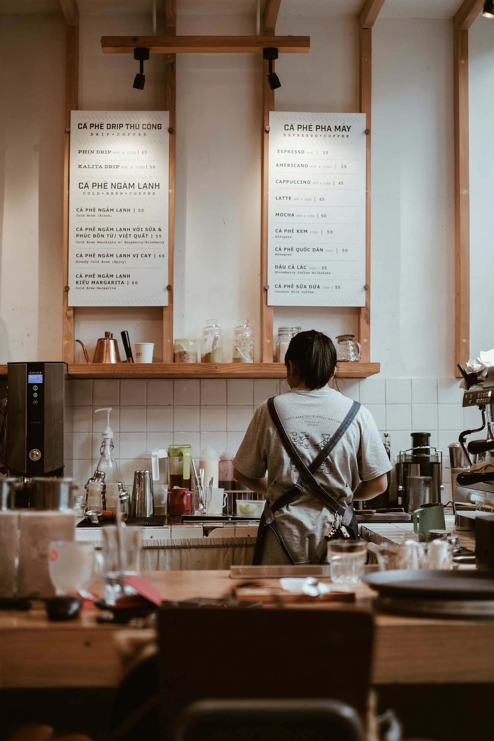 Barista de costas em frente ao quadro de menu de uma cafeteria, em ambiente real e aconchegante.