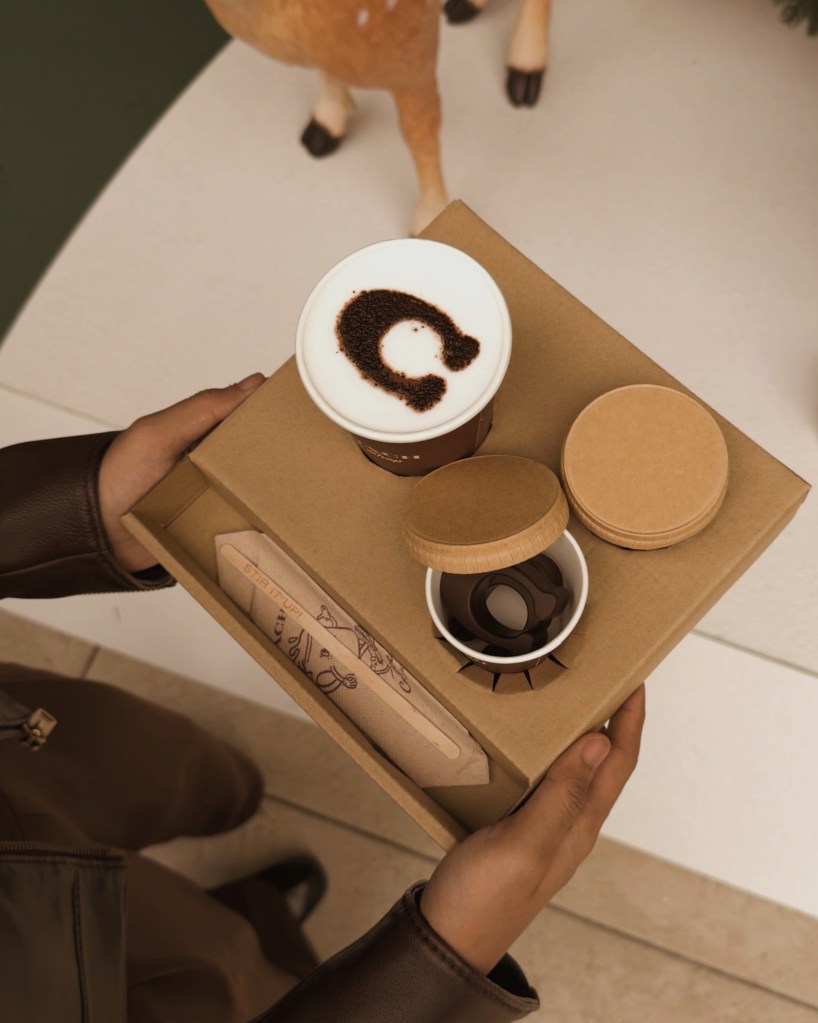 A hand holding a takeaway container with two cups of coffee, one featuring latte art, and a box containing treats, set against a festive background.