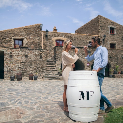 A man and woman drink wine by a white barrel in front of a rustic stone building with stairs.