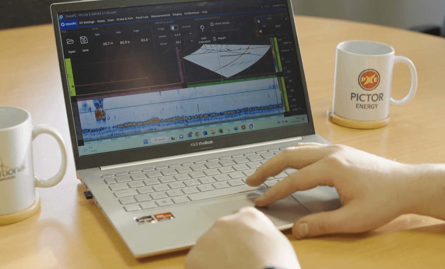 Hands on a laptop with a graph on the screen, placed on a wooden table with two branded mugs. The scene conveys focus and work environment.