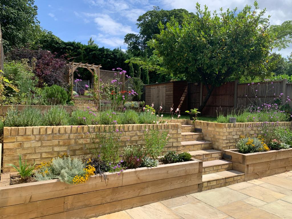 A landscaped garden featuring various plants, steps leading up, and a stone wall under a sunny blue sky.