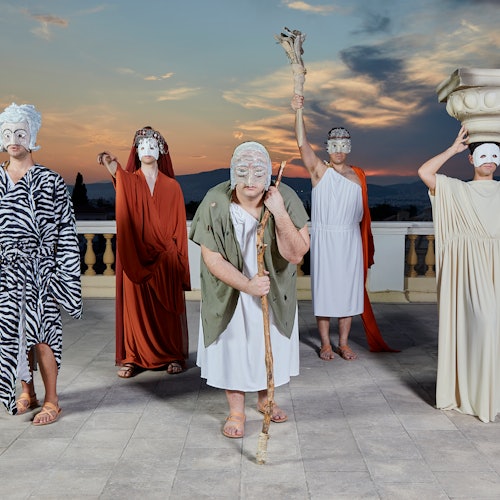 五位身着古希腊风格服装并佩戴面具的人站在屋顶上,背景是群山和壮丽的落日。