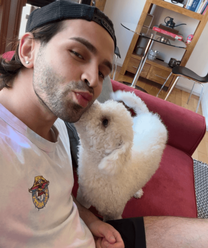 A person wearing a white shirt and a cap smiles affectionately on a red couch as a fluffy white dog approaches their face, with a wooden bookshelf and a small table in the background.