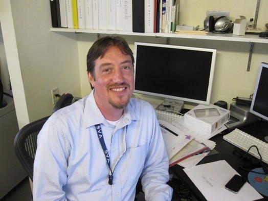 A photo of a man in a shirt sitting at a desk near his computer.