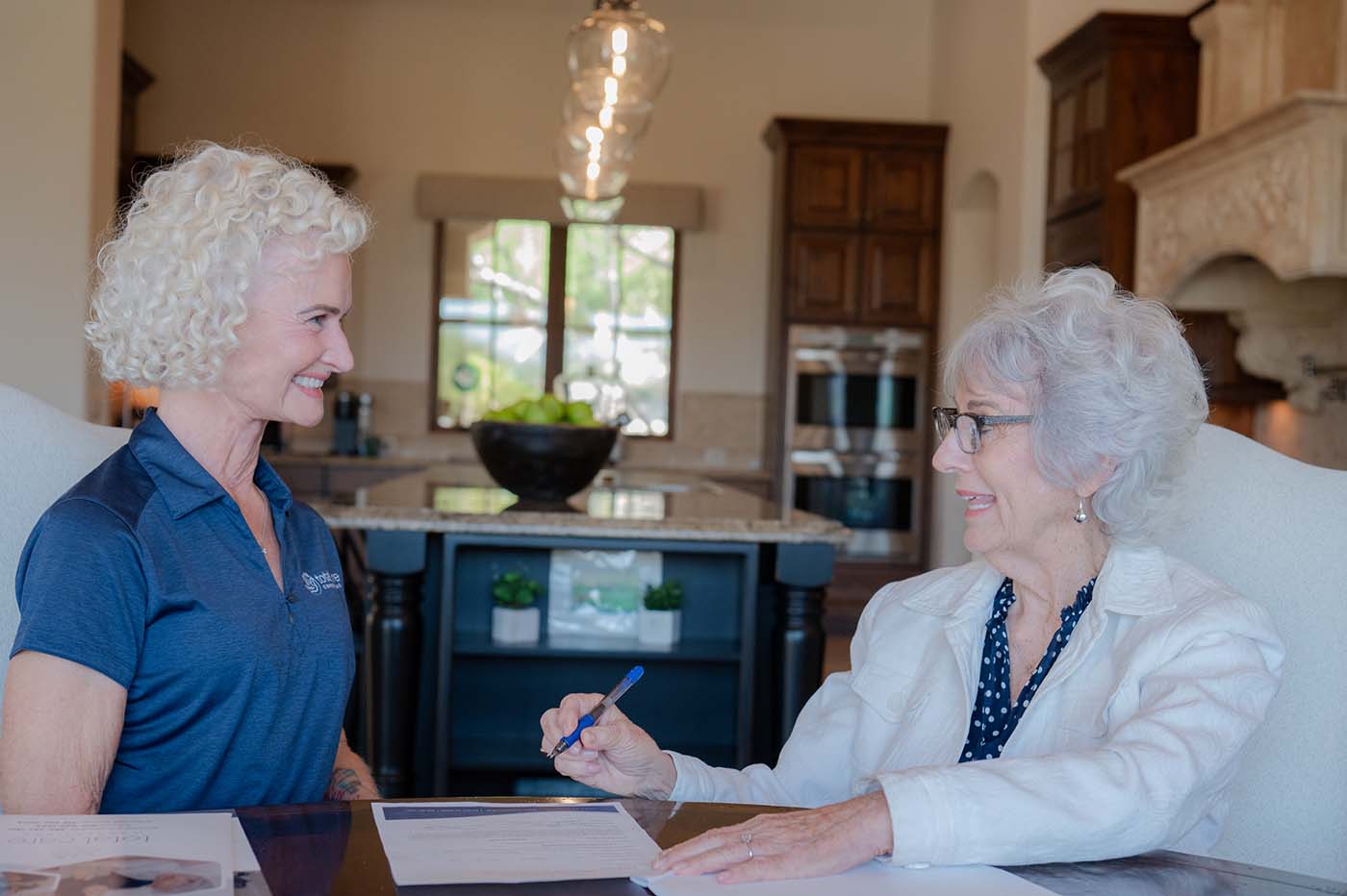 Two women sitting at a table smiling at each other, one holding a pen over a document.