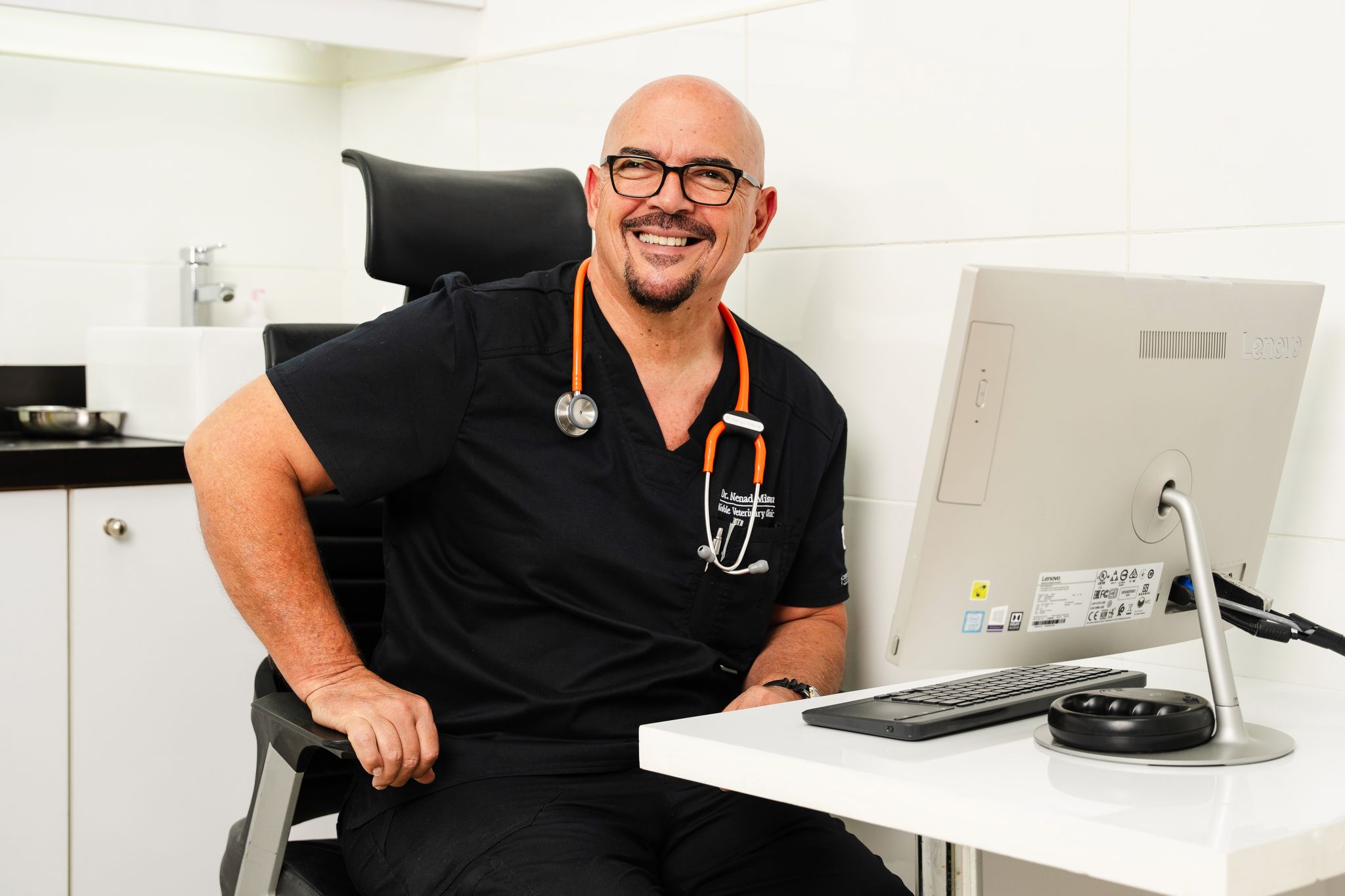 A smiling male veterinarian wearing black scrubs and an orange stethoscope sits at his desk at Noble Vet Clinics, ready to assist pet patients.