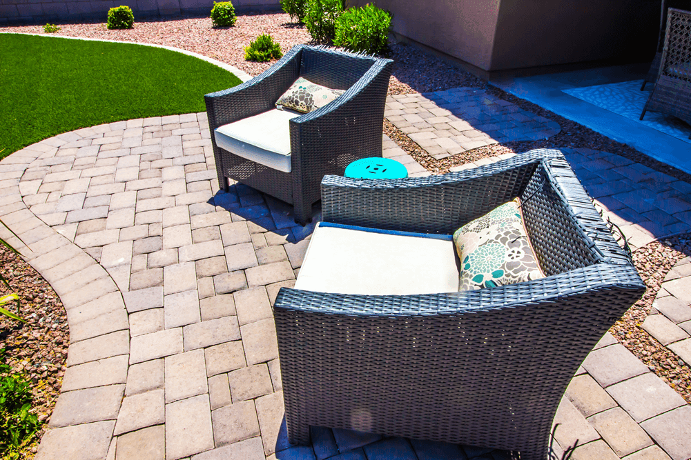 Two wicker chairs with cream cushions and floral throw pillows placed on a curved paver patio. The scene is surrounded by neat landscaping, featuring artificial grass and a mix of stone and mulch.