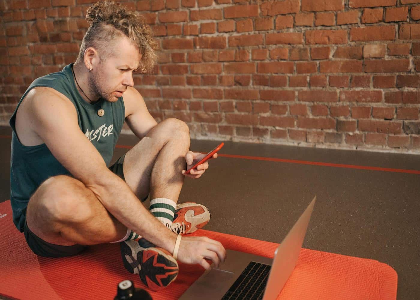 Man sitting on orange yoga mat with laptop in front of him looking at smartphone