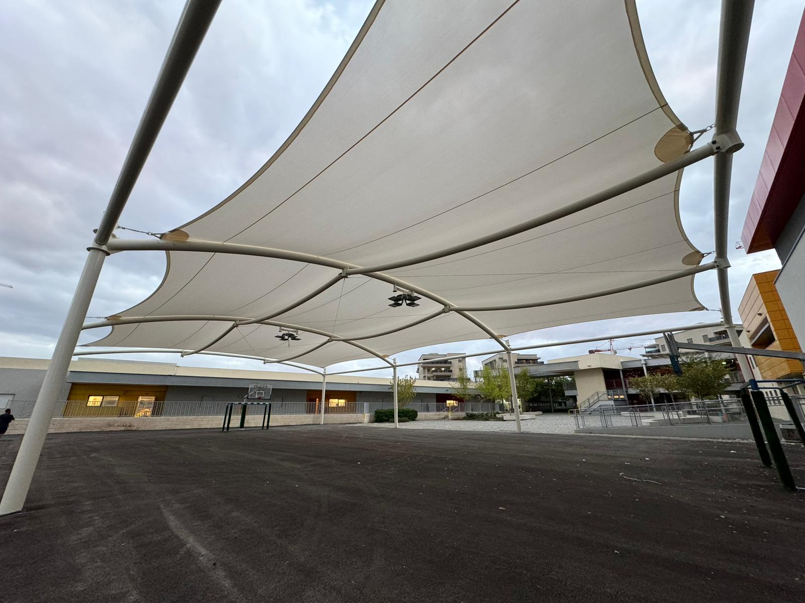 Large beige shade sails over an empty schoolyard with a basketball hoop and buildings in the background.