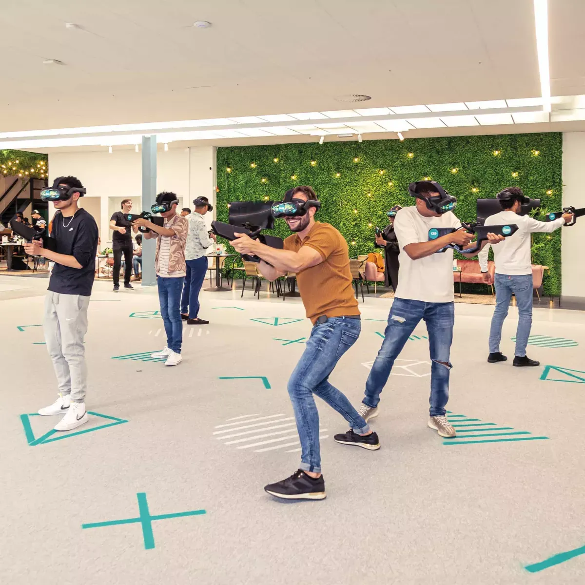 Multiple players wearing VR headsets and haptic vests moving freely across a large indoor arena floor, with a digital projection of the Cape Town skyline in the background.