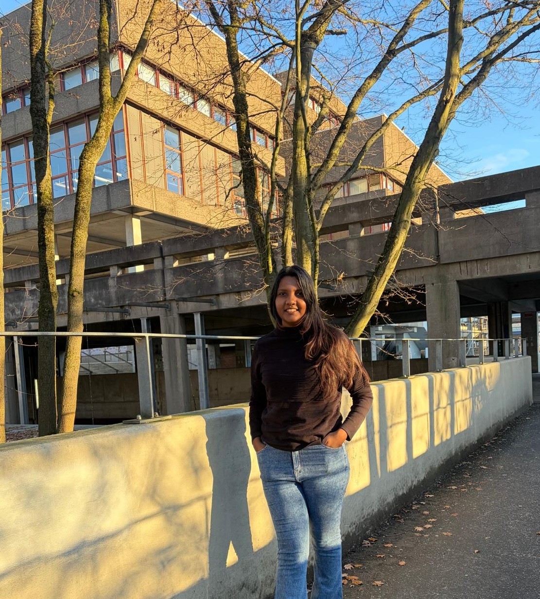 Smiling young woman with long hair standing against a dark green background, holding a finger to her chin.
