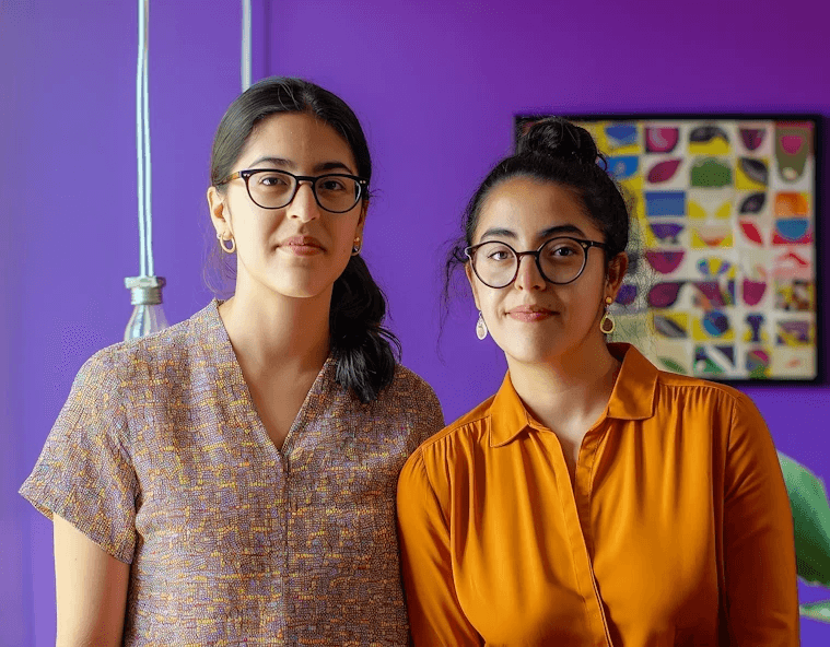 Two women with glasses stand against a vibrant purple wall, wearing patterned and orange blouses, with a colorful painting in the background.