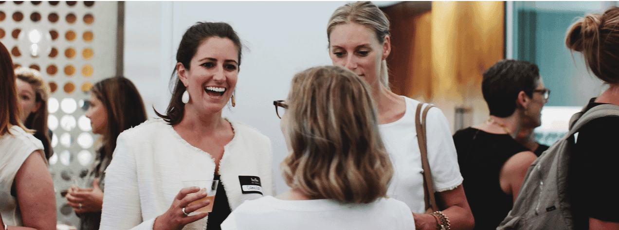 A group of three women engaged in conversation at a social event, with people milling about in the background.