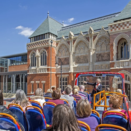 Touristen sitzen in einem offenen Doppeldeckerbus und lauschen einem Reiseführer vor einem reich verzierten, historischen Gebäude unter einem klaren blauen Himmel.
