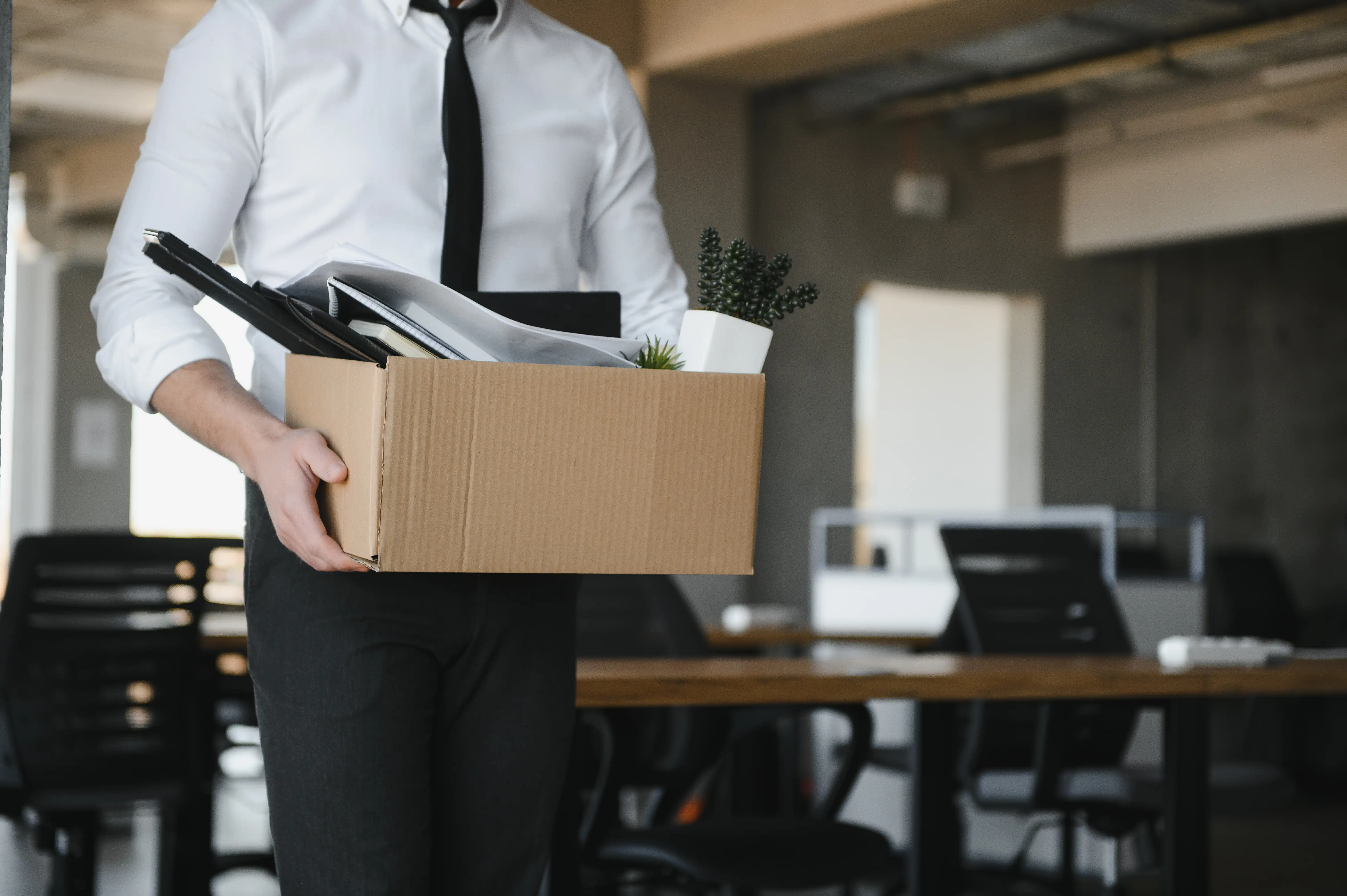 Professional worker carrying cardboard box of personal office belongings after losing a job, illustrating the impact of workplace relocation fraud in the Central Valley