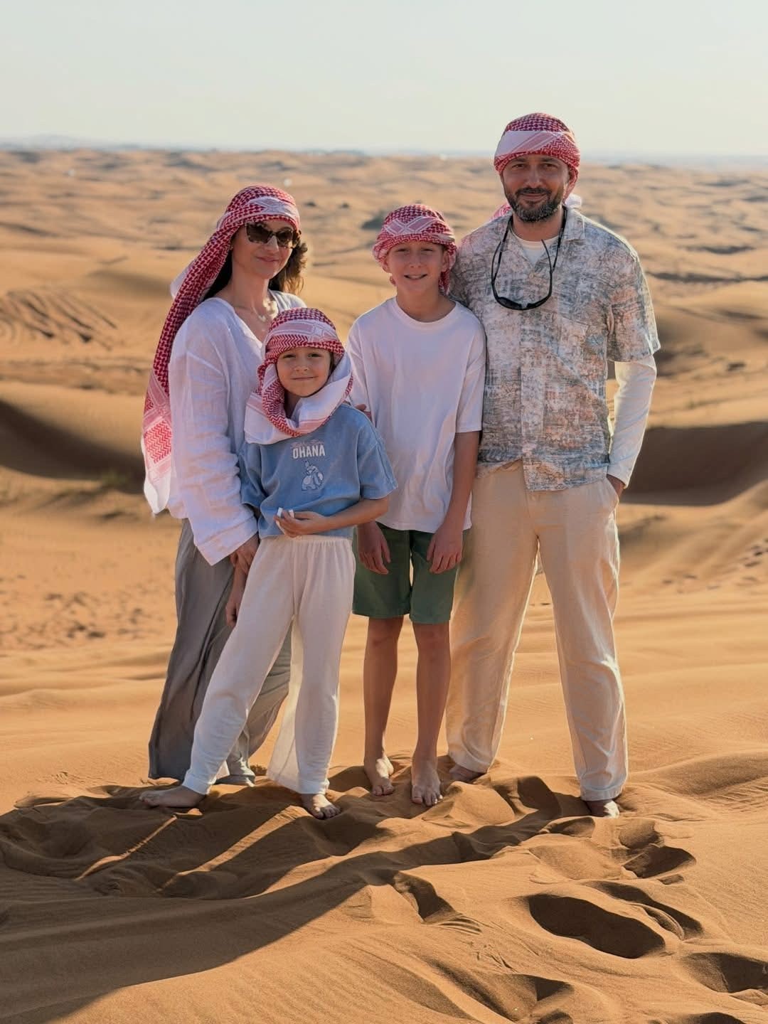 A family of four wearing traditional red and white keffiyehs, smiling together on the golden sand dunes during a private Dubai desert safari