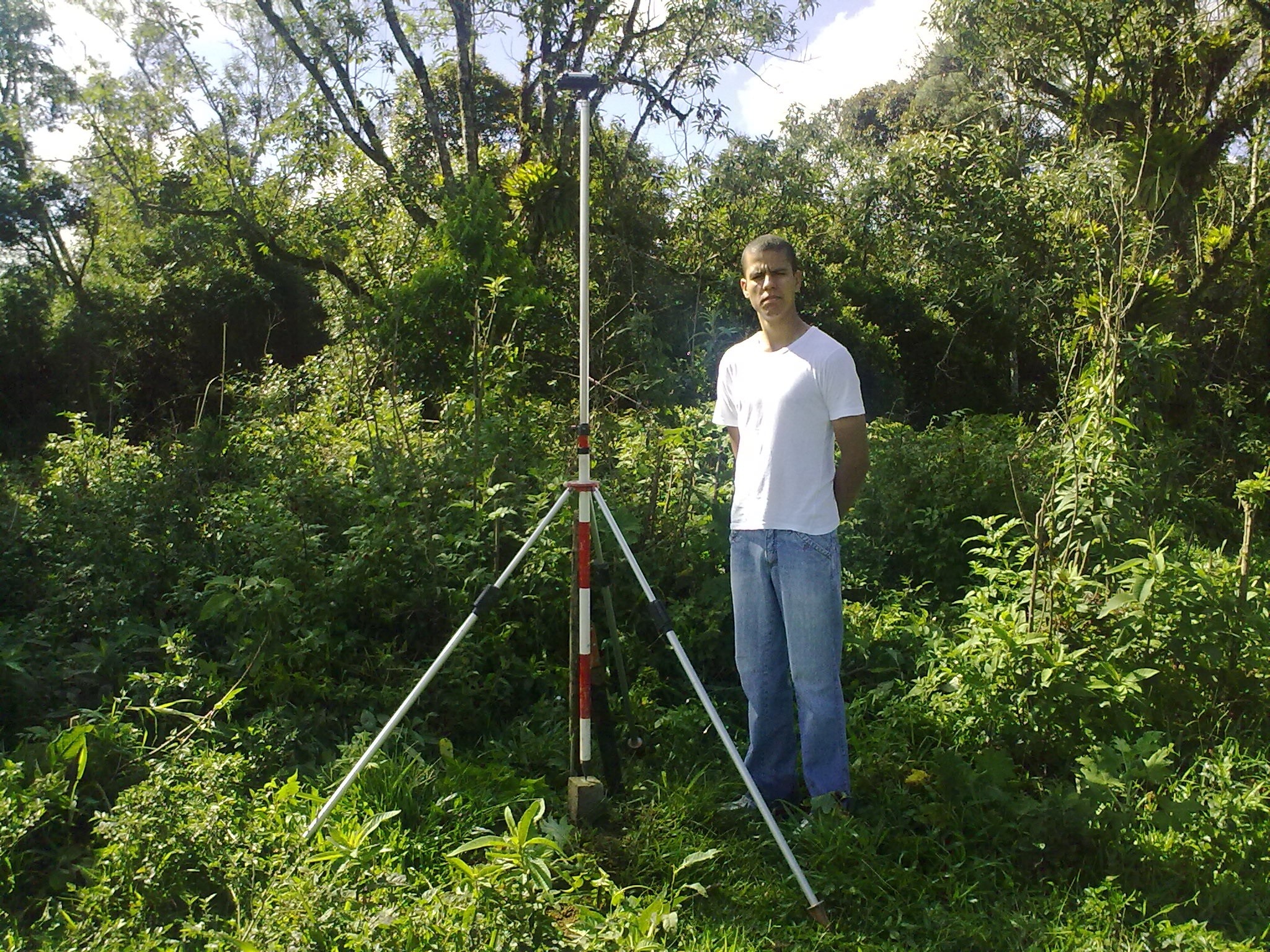 Profissional realizando levantamento topográfico em campo com bastão e prisma sobre tripé, cercado por vegetação densa e árvores.
