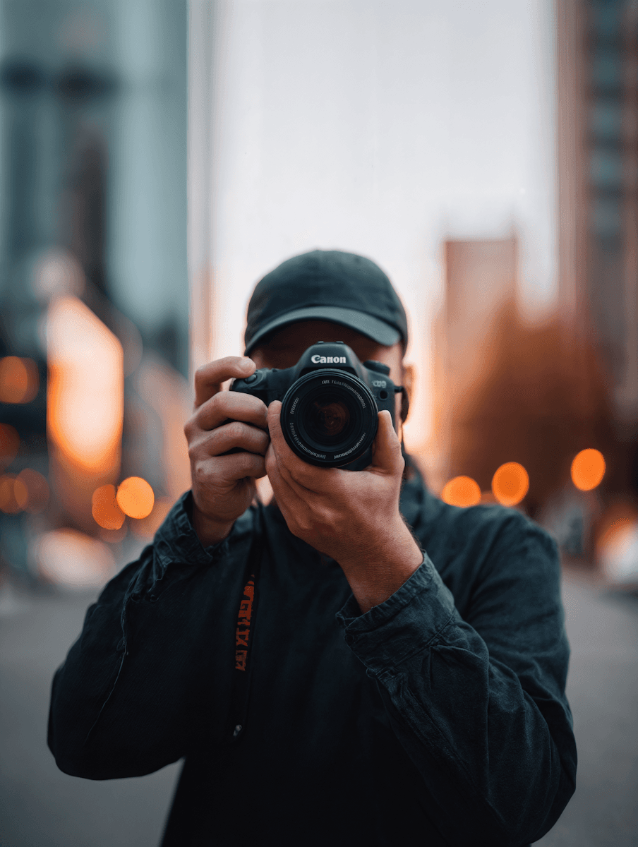 A man wearing a black cap taking a photo with a Canon camera in an urban area with bokeh effects.