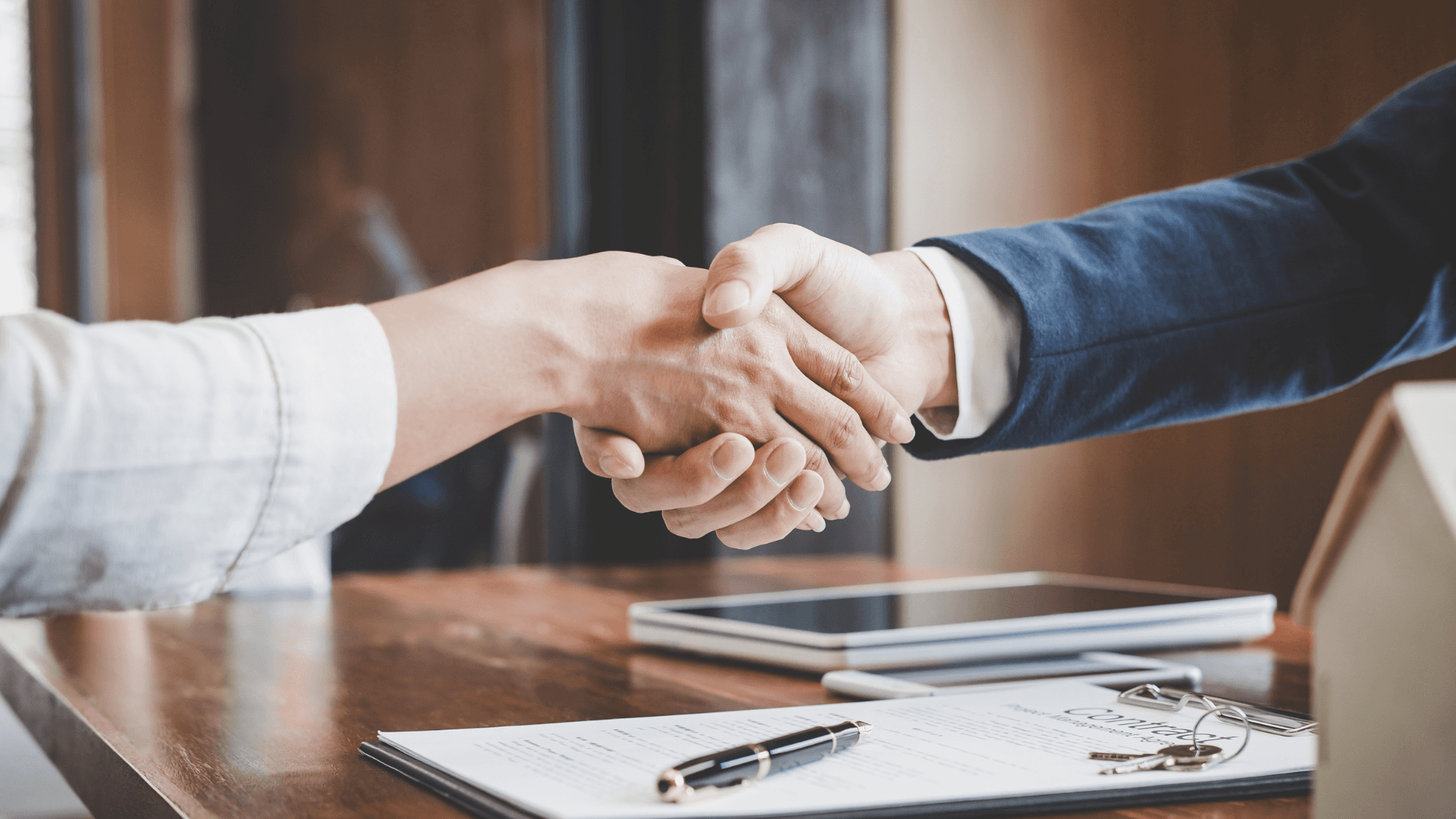 Zwei Personen schütteln sich über einem Tisch die Hände und symbolisieren eine Vereinbarung oder Partnerschaft in einem professionellen Umfeld.