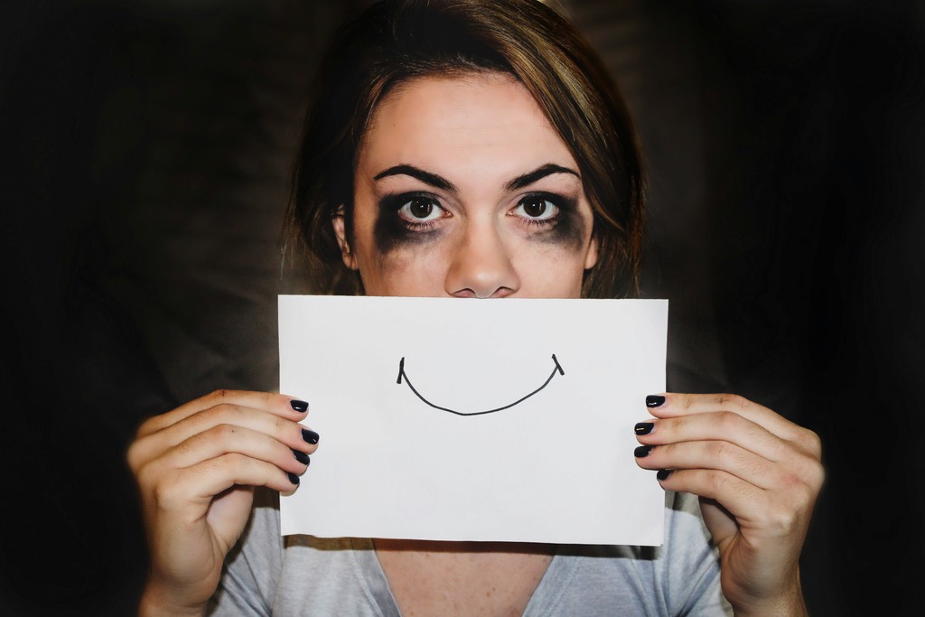 A woman with smudged makeup holds up a piece of paper with a drawn smile, conveying the struggle of hiding inner pain behind a facade.