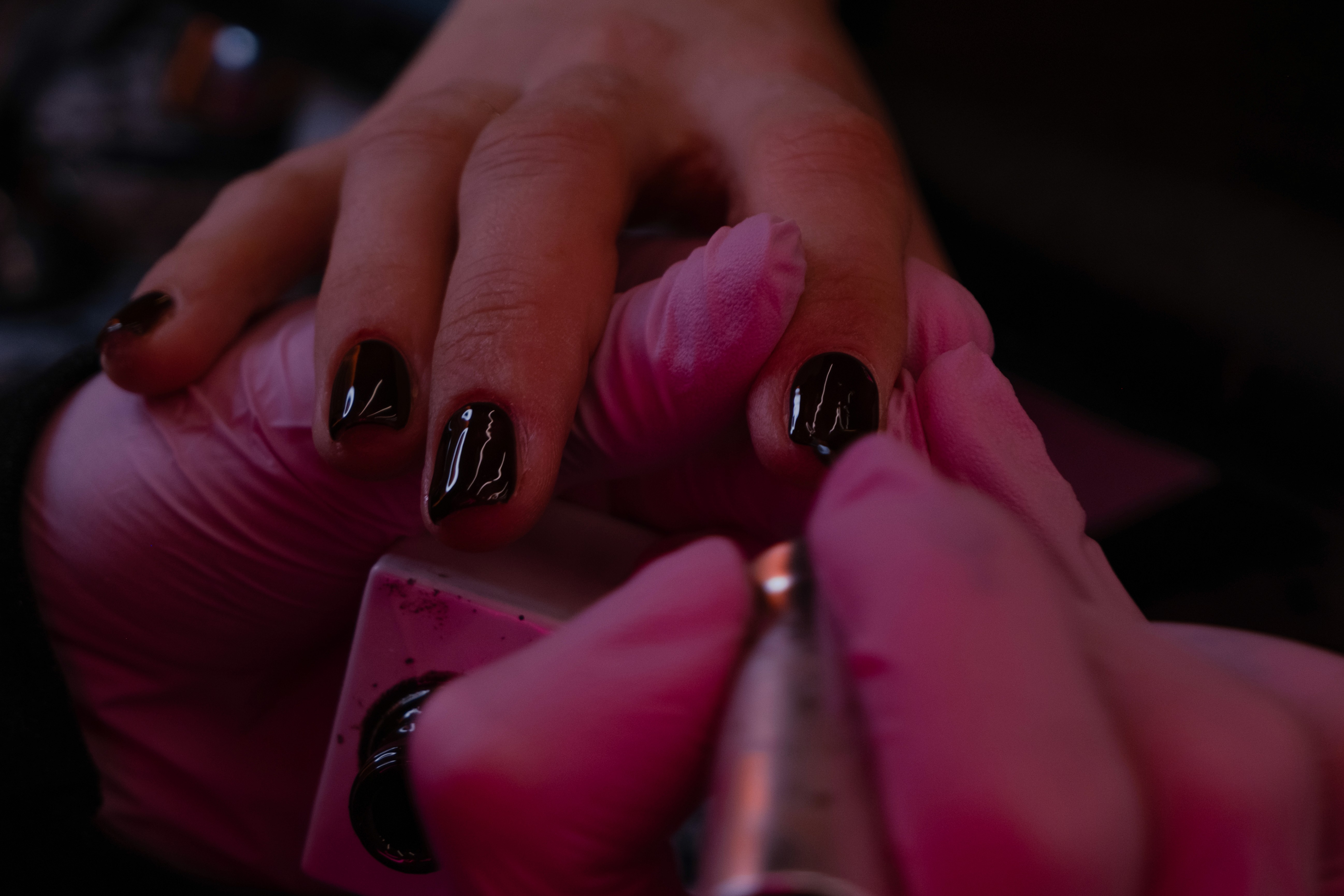 A close-up, moody shot of a technician in pink nitrile gloves carefully applying a glossy black gel polish to a client’s short, natural nails.