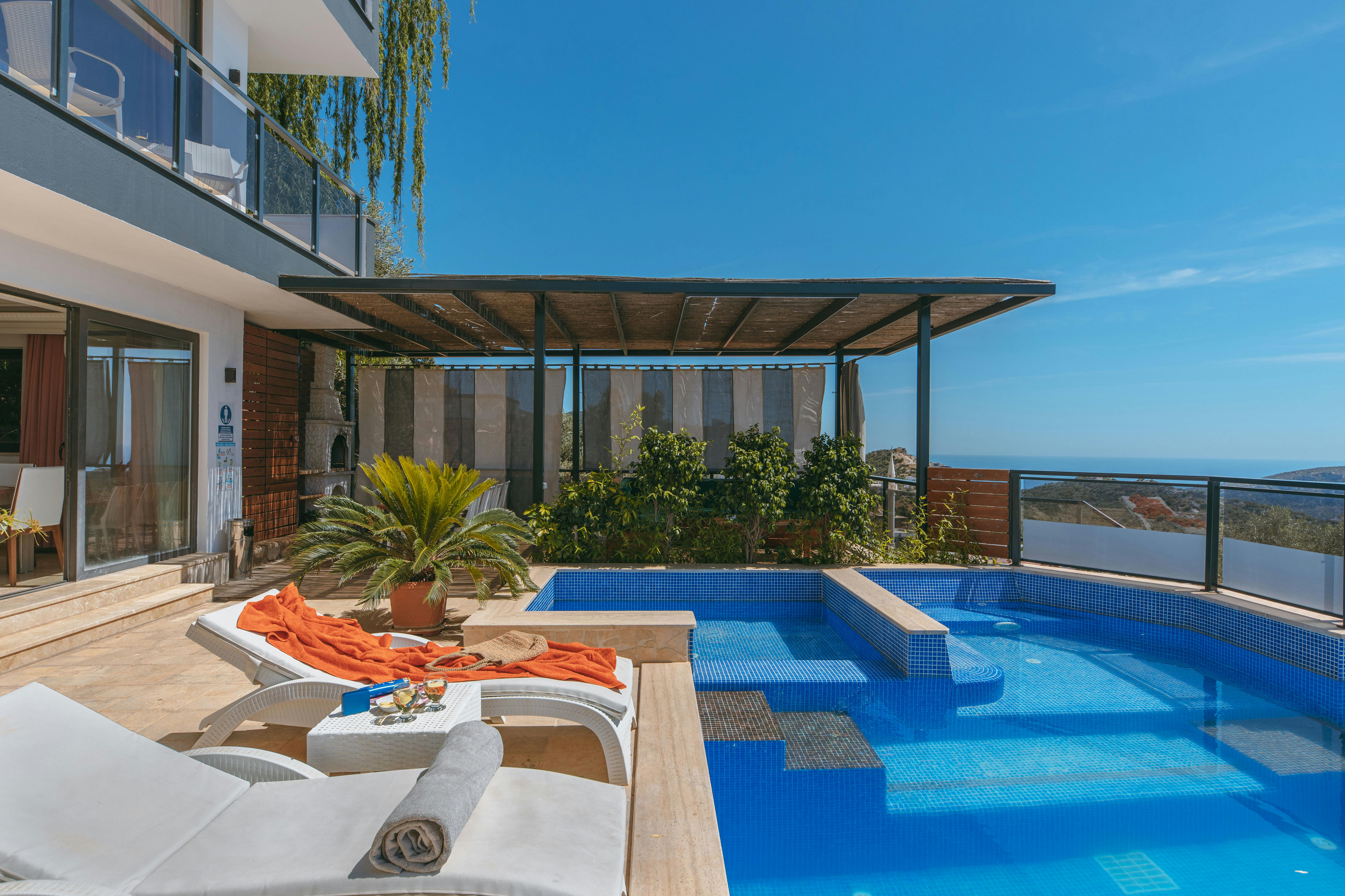 A luxurious outdoor poolside deck features modern lounge chairs with bright orange towels, surrounded by lush greenery and overlooking a scenic ocean view under a clear blue sky.