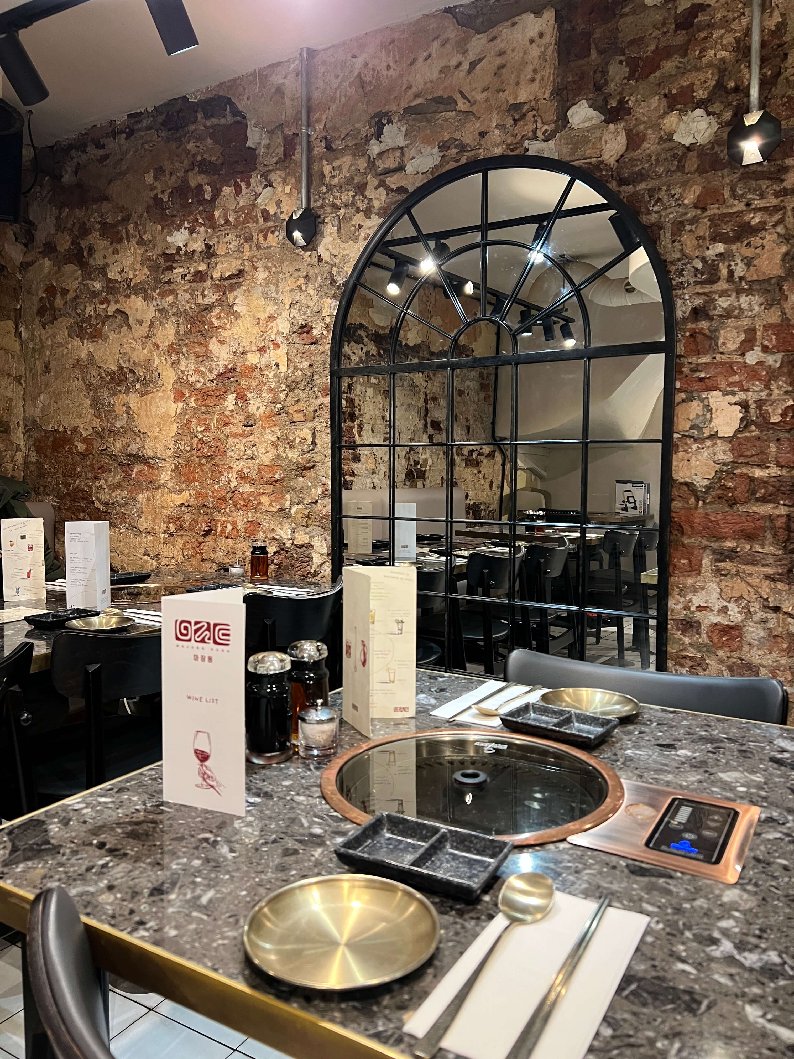 Cozy cafe corner with a round terrazzo table surrounded by wooden chairs, decorated with potted plants and a large mirror reflecting warm light.
