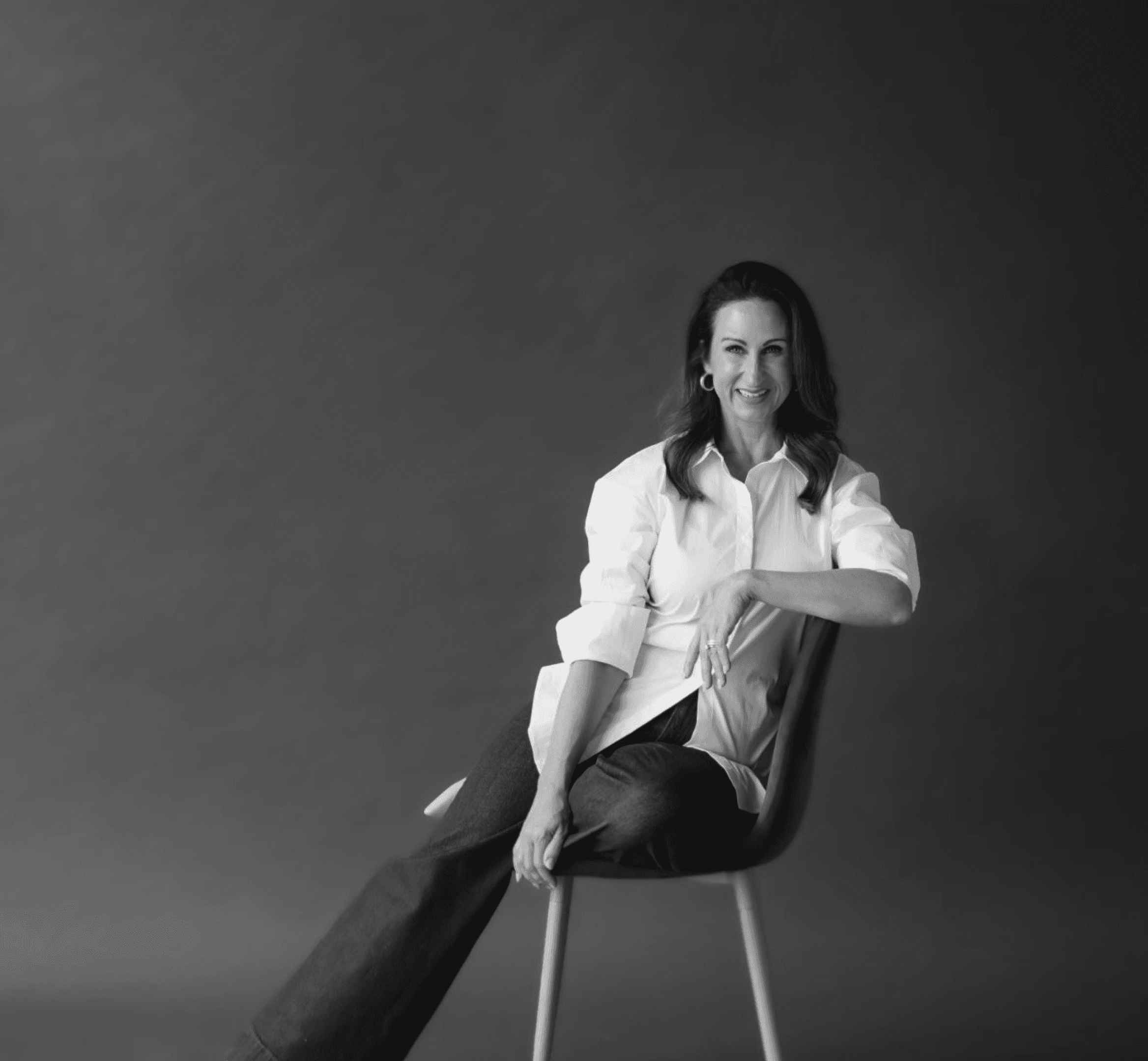 Stacy sits relaxed on a chair, resting one foot on the seat, smiling confidently at the camera.