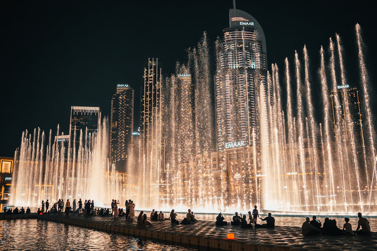The Dubai Fountain Show at night, with tall golden water jets shooting up in front of the skyscrapers and people watching.&nbsp;