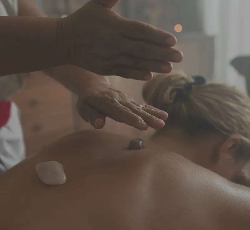 Yäan Spa en Be Tulum: mujer recibiendo un relajante masaje con dos piedras calientes colocadas sobre ella, mostrando las manos de la masajista.