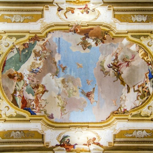 Barok plafondfresco met figuren en wolken, omringd door verfijnde goud- en witgekleurde architectonische details.