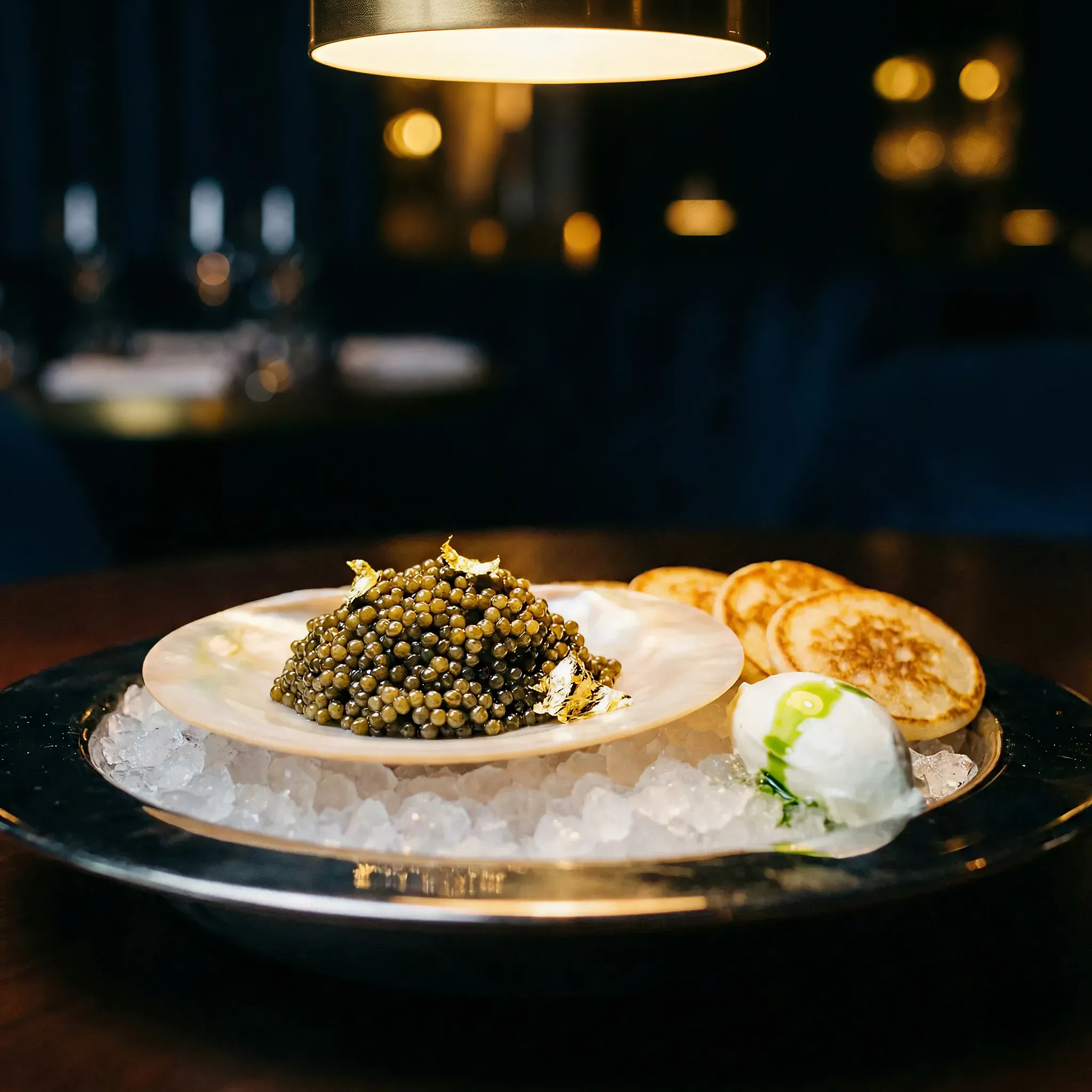 A lavish dish of caviar rests on a bed of ice, surrounded by blinis and a dollop of cream. A warm light casts a cozy glow in an elegant setting.