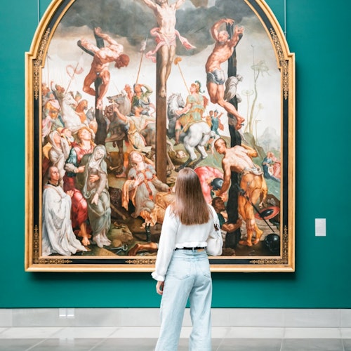 一位长发女子站在画廊的青绿色墙壁前，凝视一幅大型细致的十字架画作。