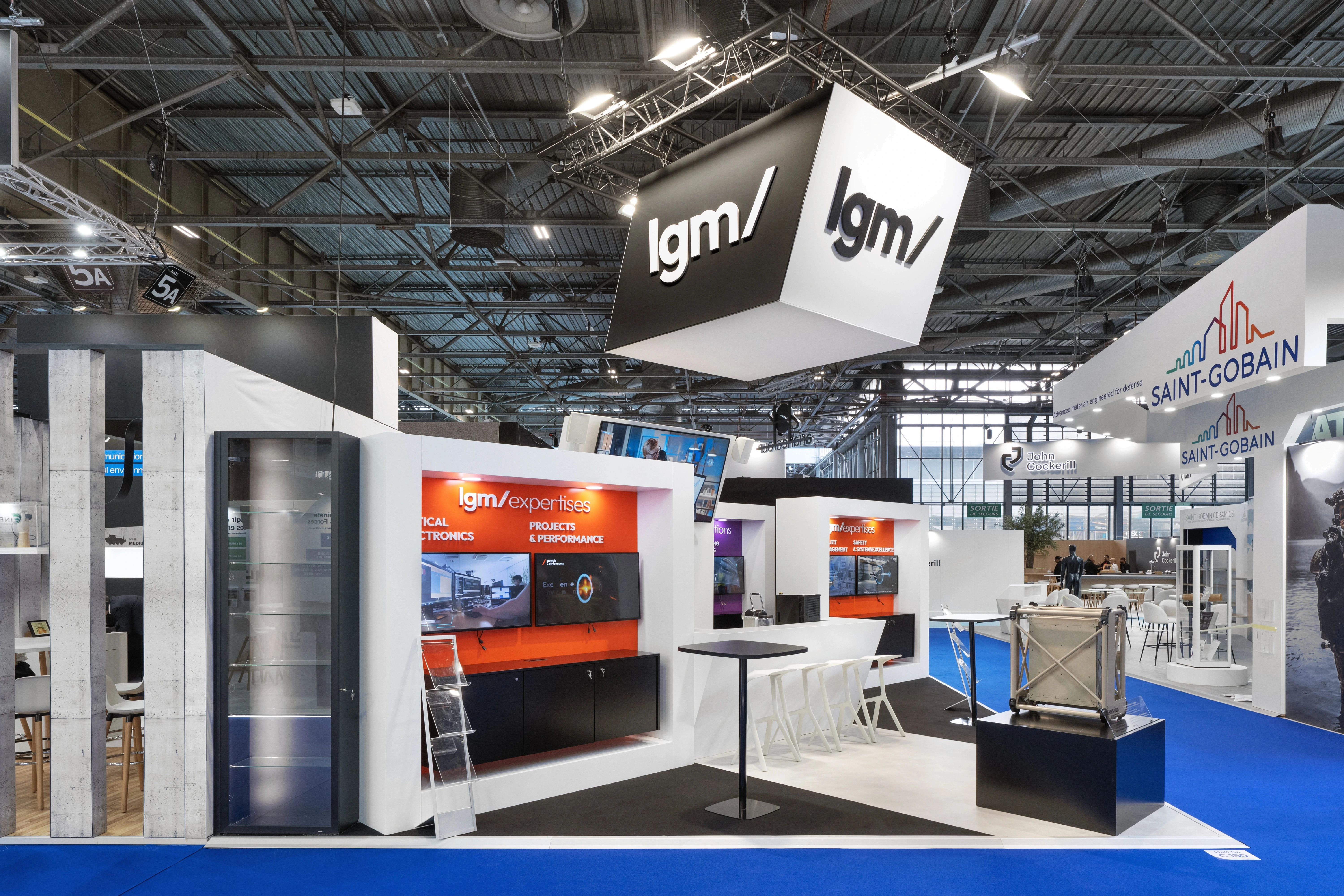 Stand LGM ingénierie de défense conçu par EXPACE à Eurosatory 2026, Hall 5A — cube suspendu lgm/ noir et blanc, pôles expertises orange et transformations violet