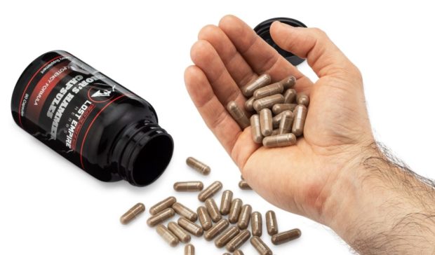 A hand holds several capsules above a bottle, with more capsules scattered around. The bottle is black with a label.