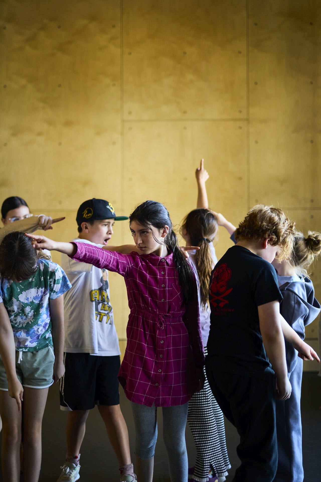 A group of young performers stand closely together in a rehearsal room, each extending an arm in a different direction as part of a movement exercise.