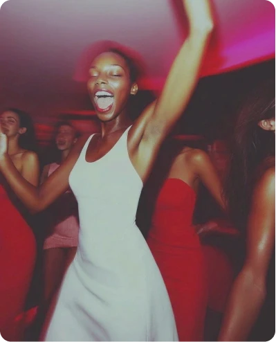 A group of young women celebrating and dancing at a party with red ambient lighting, featuring a woman in a white dress in the foreground