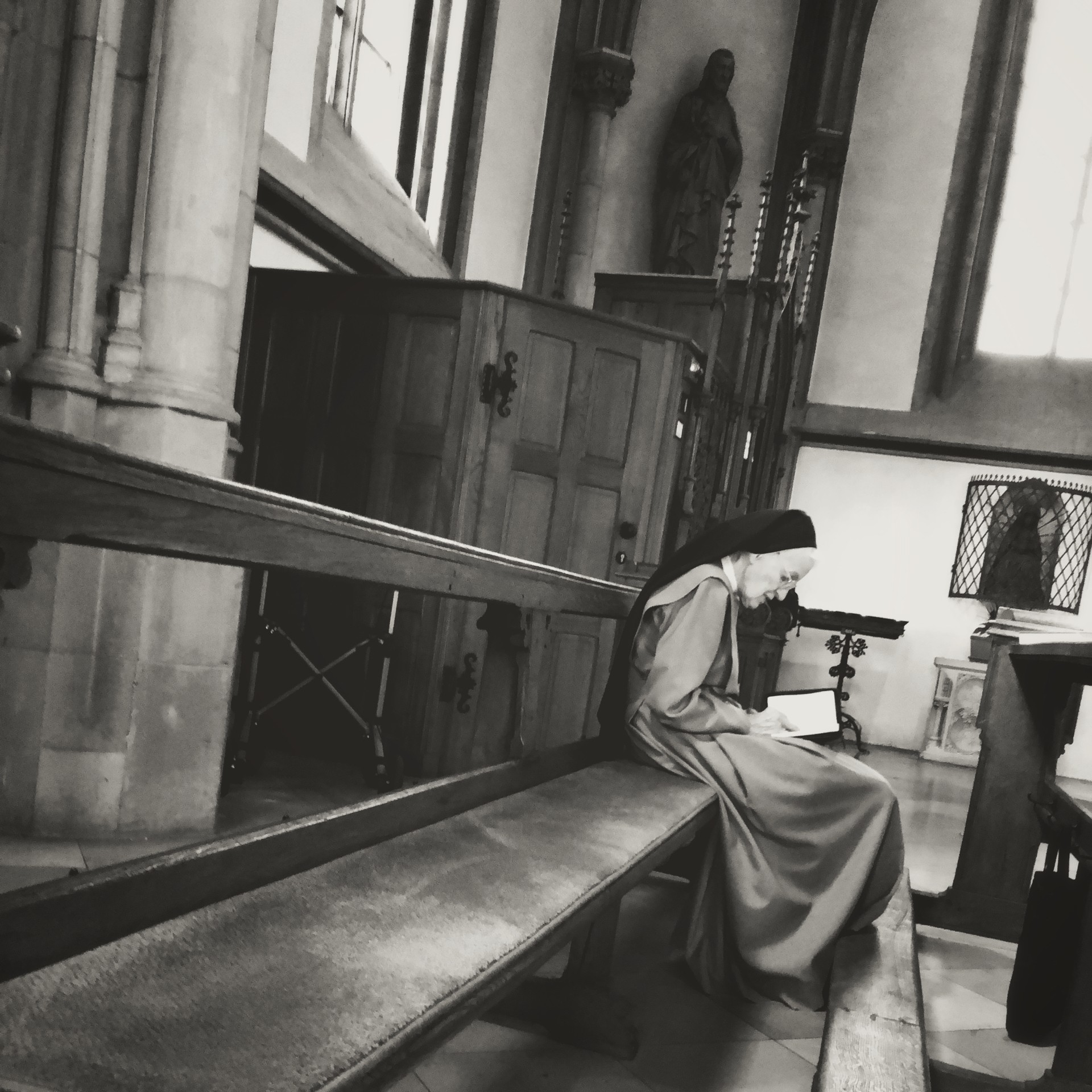 Nun sitting on a long wooden bench in a church, wearing traditional religious attire and a hat.