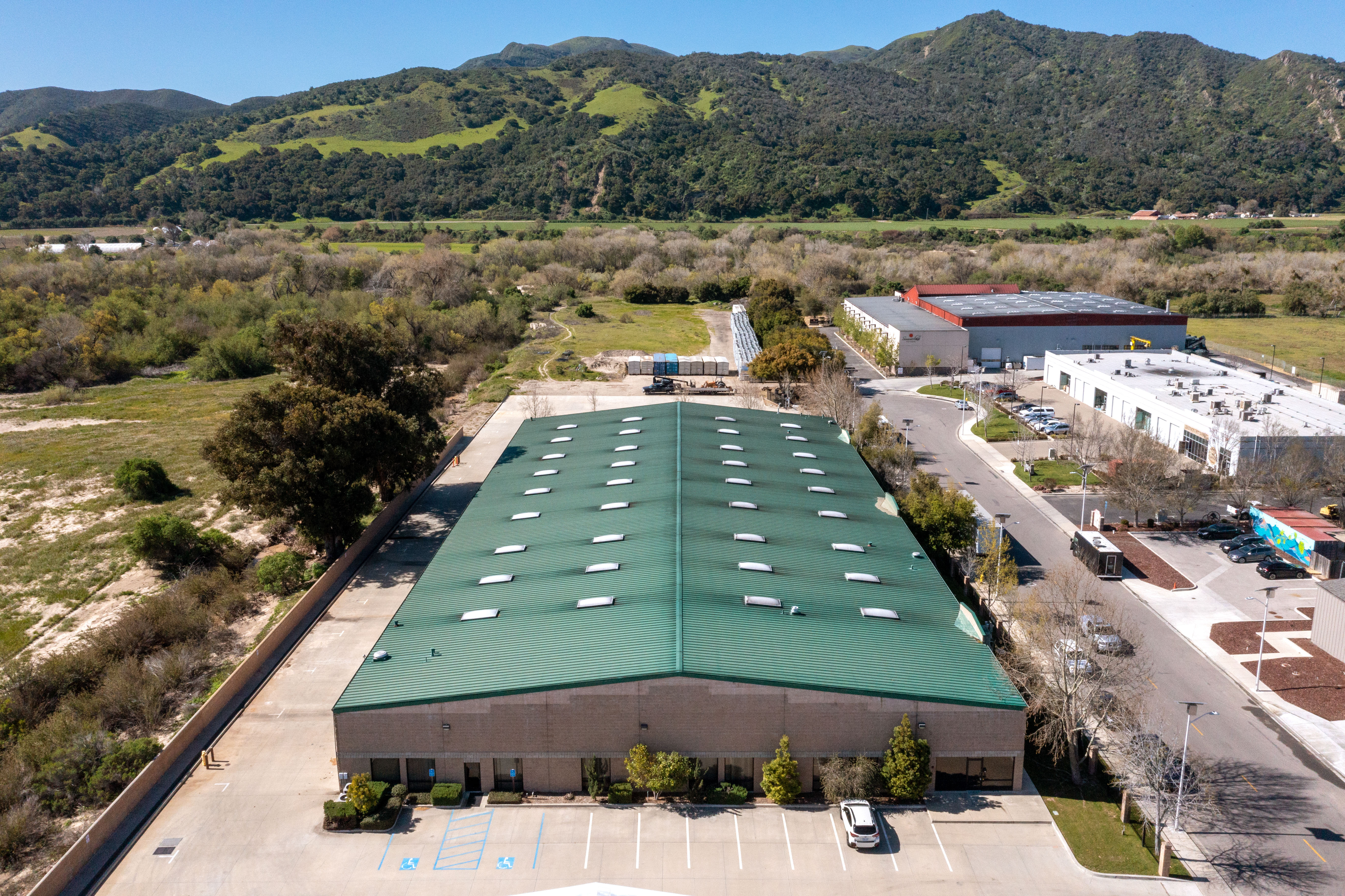 Aerial view of a large single-story warehouse or storage building with a green metal roof, skylights, and a paved parking lot, situated on a flat parcel bordered by open scrubland and green mountain foothills under a clear blue sky.
