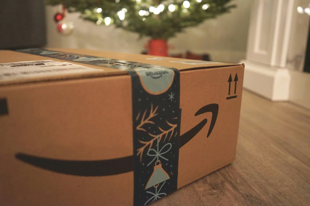 A close-up view of a delivery box with a recognizable logo, festively adorned with a snowflake design, positioned on a floor with a blurred Christmas tree in the background, evoking the holiday spirit.